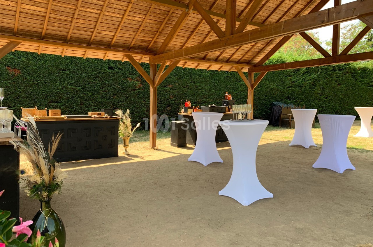 Espace extérieur couvert avec tables hautes drapées de blanc, buffet et décoration végétale sous une structure en bois.
