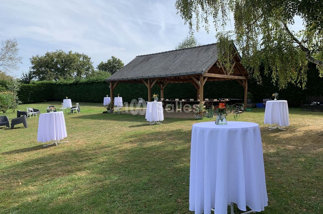 Tables hautes dressées avec nappes blanches dans un jardin, près d'un abri en bois, sous un ciel dégagé.