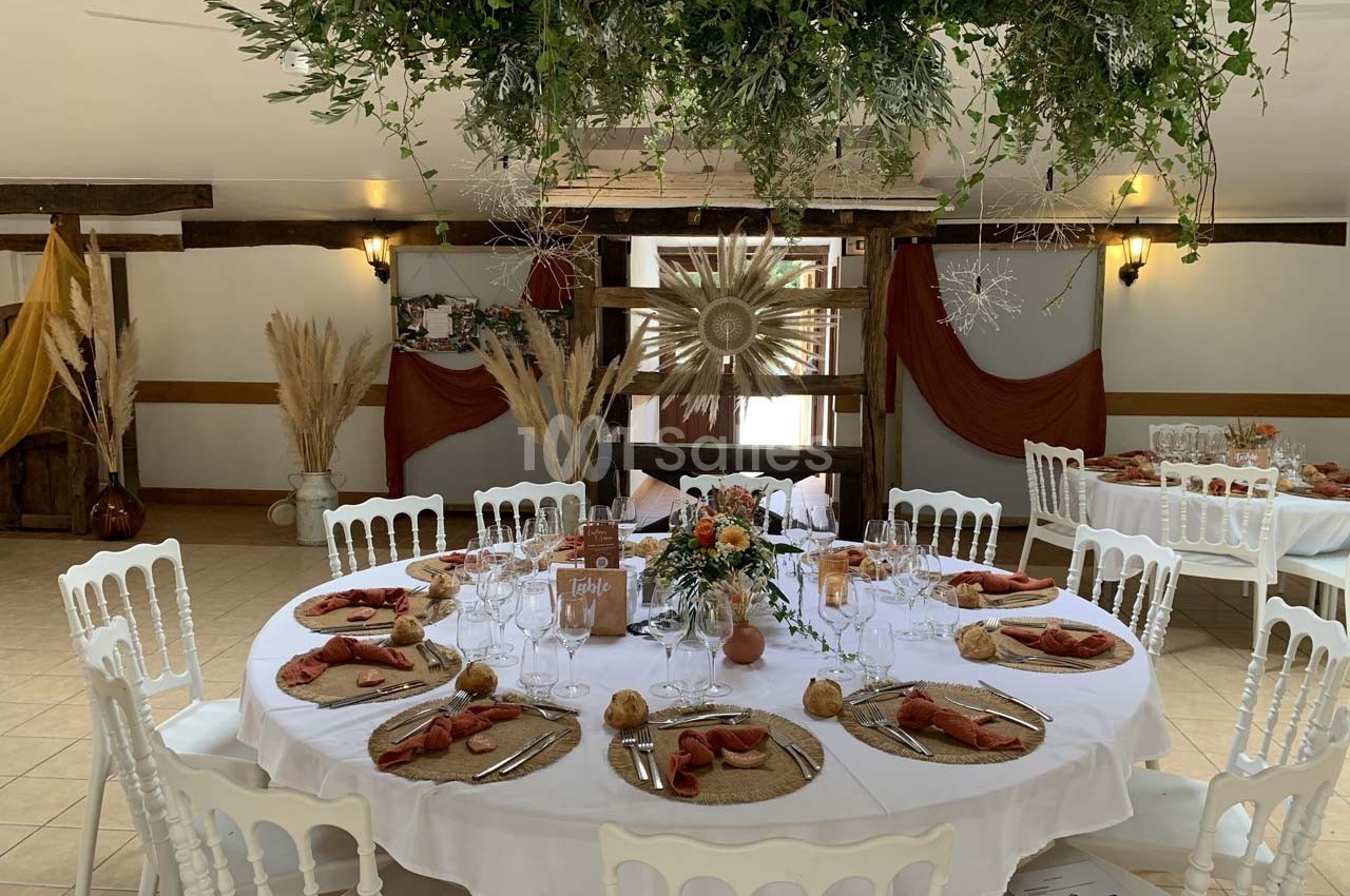 Table ronde décorée pour un événement, avec nappes blanches, serviettes orange, vaisselle élégante et centre de table floral.