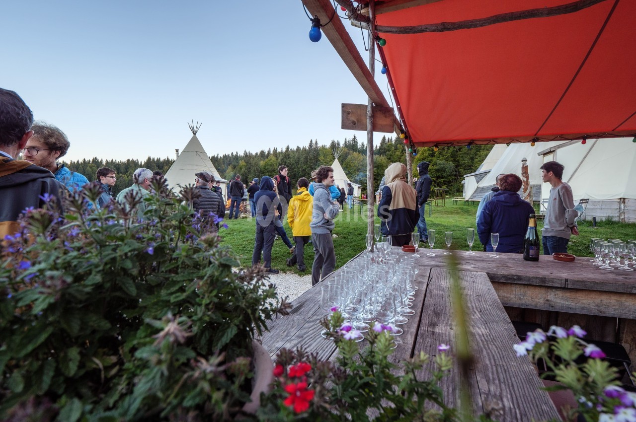 Groupe de personnes rassemblées en extérieur près de tipis, avec une table garnie de verres au premier plan.