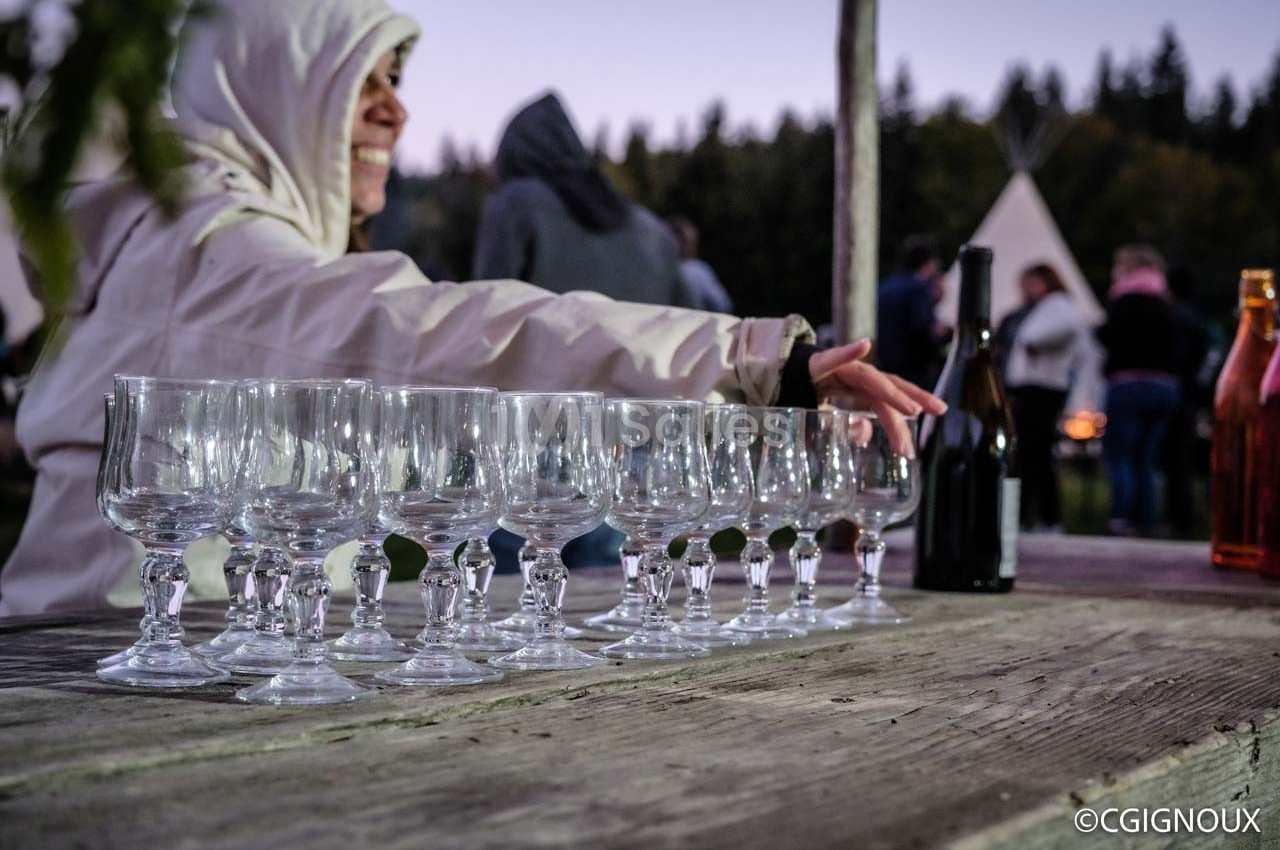 Une personne en veste claire dispose des verres sur une table en bois, avec un paysage extérieur en arrière-plan.