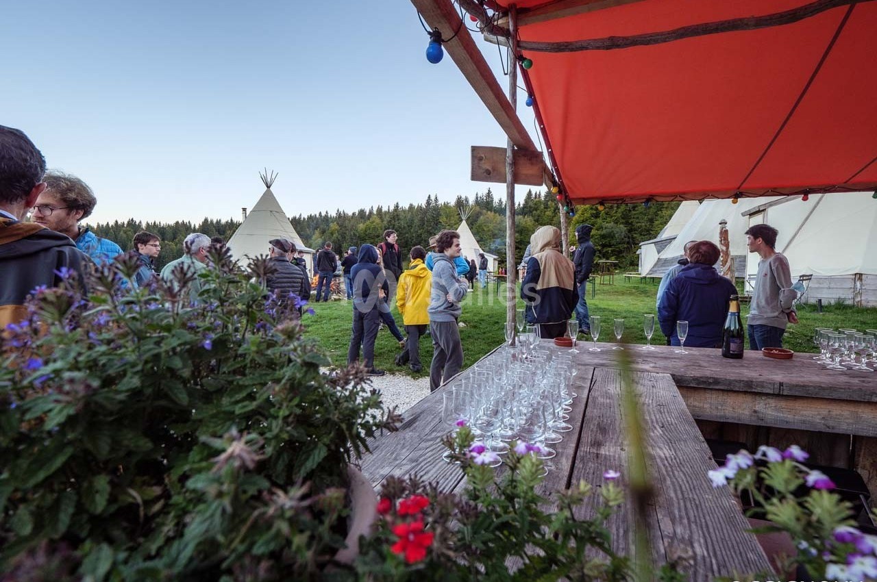 Groupe de personnes rassemblées en extérieur près de tentes et d'une table avec des verres, dans un cadre naturel.