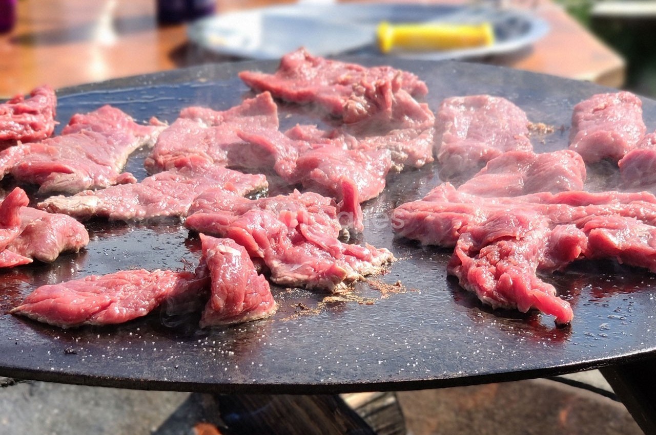 Viande rouge en train de griller sur une plancha en extérieur, avec des ustensiles visibles en arrière-plan.