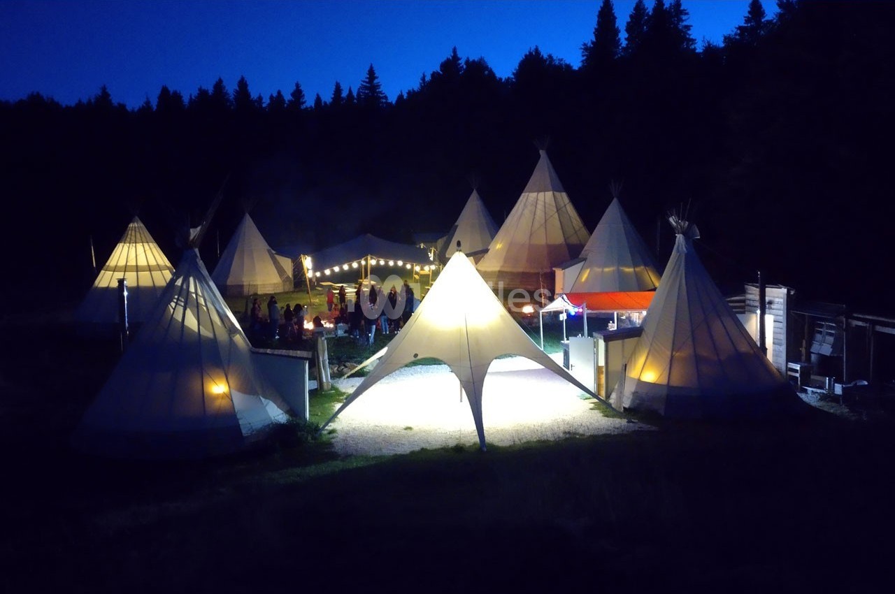 Campement de tipis éclairés la nuit, entouré de forêt, avec des lumières et des personnes rassemblées.