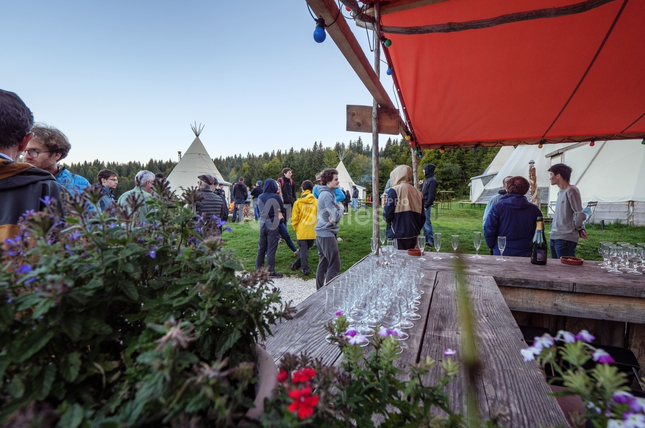 Groupe de personnes rassemblées en extérieur près de tipis, avec une table garnie de verres au premier plan.