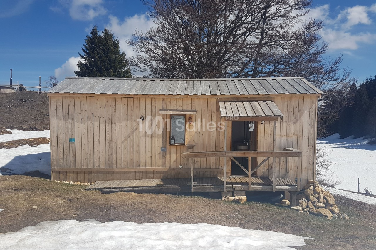 Cabane en bois sur un terrain partiellement enneigé, entourée d'arbres et sous un ciel bleu.