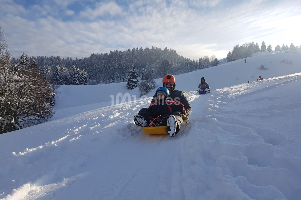Deux personnes descendent une pente enneigée en luge, entourées d'un paysage de forêt sous un ciel partiellement dégagé.