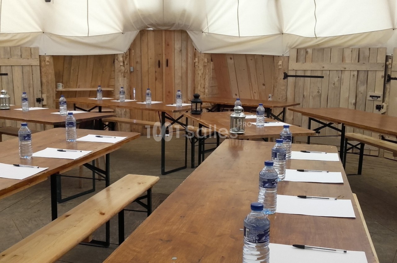 Tables en bois disposées en U avec bancs, bouteilles d'eau, blocs-notes et stylos dans une salle à structure en toile.