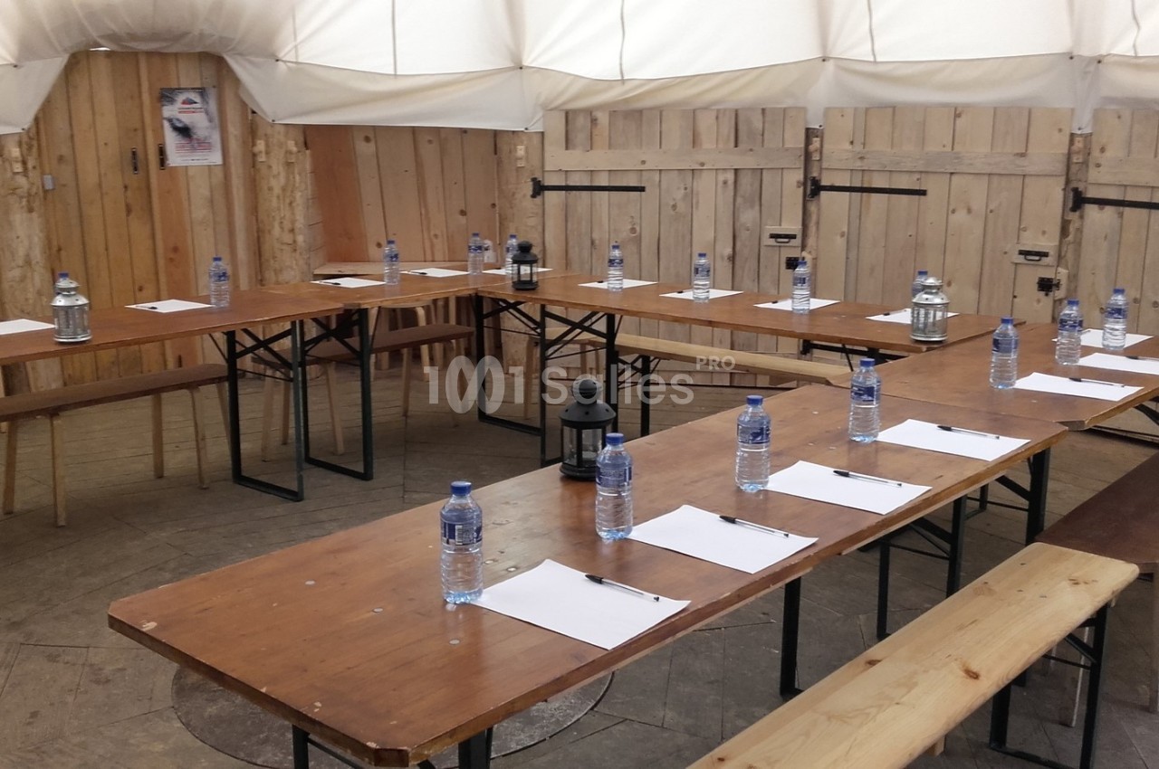 Disposition en U de tables en bois avec bouteilles d'eau, blocs-notes et bancs dans une salle lumineuse au style rustique.