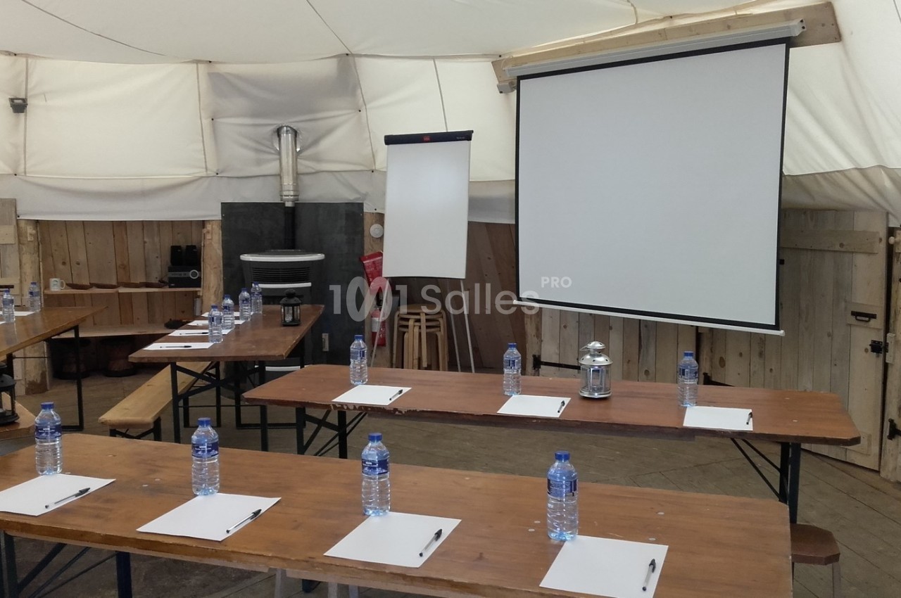 Salle de réunion rustique avec tables en bois, bouteilles d'eau, blocs-notes et écran de projection.