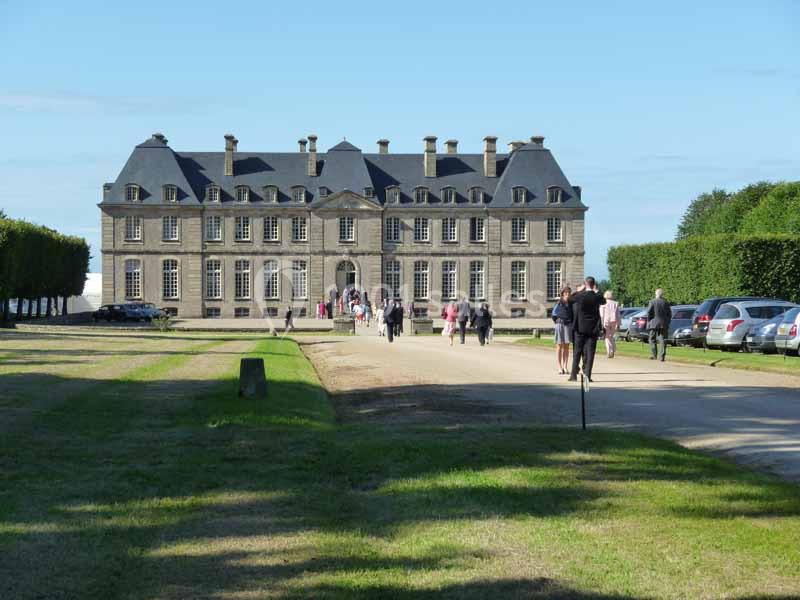 Façade d'un château en pierre entouré de pelouses, avec des visiteurs marchant sur une allée gravillonnée.
