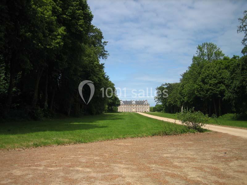 Allée bordée d'arbres menant à un grand bâtiment en pierre sous un ciel partiellement nuageux.