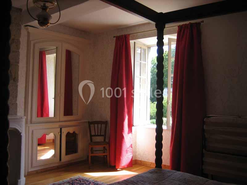 Chambre lumineuse avec lit à baldaquin, armoire à miroir, chaise en bois et rideaux rouges encadrant une fenêtre ouverte.