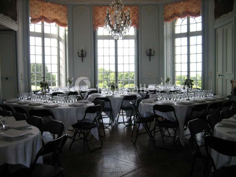 Salle élégante avec tables rondes dressées, nappes blanches, chaises noires et grandes fenêtres donnant sur un jardin.