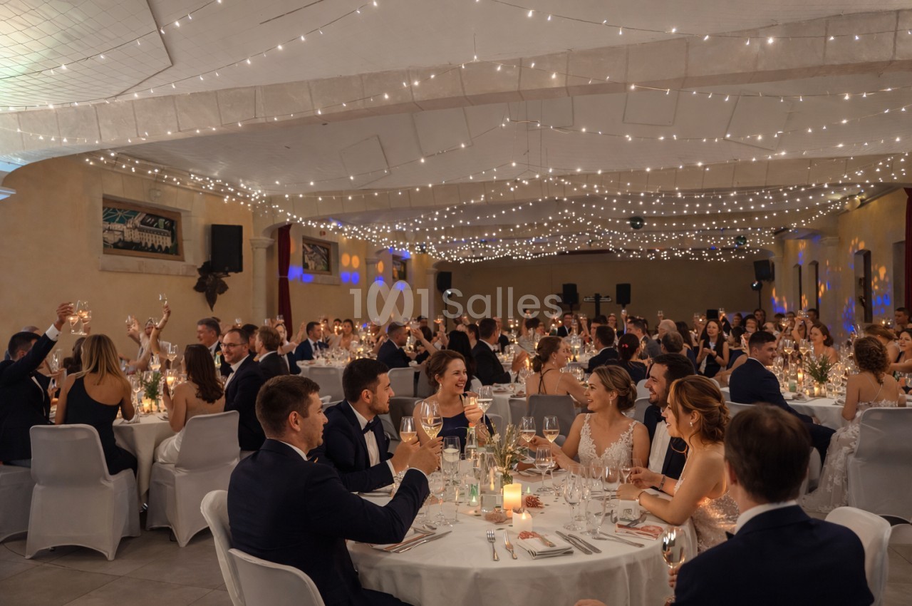 Salle de réception décorée avec des guirlandes lumineuses, des invités assis à des tables festives levant leurs verres.