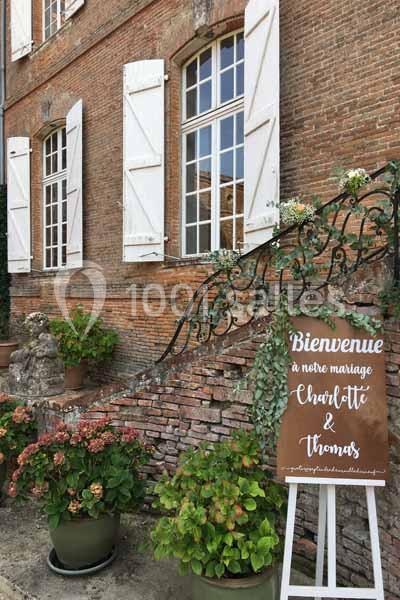 Panneau de bienvenue pour un mariage devant un mur en briques avec des fenêtres, plantes et décorations florales.