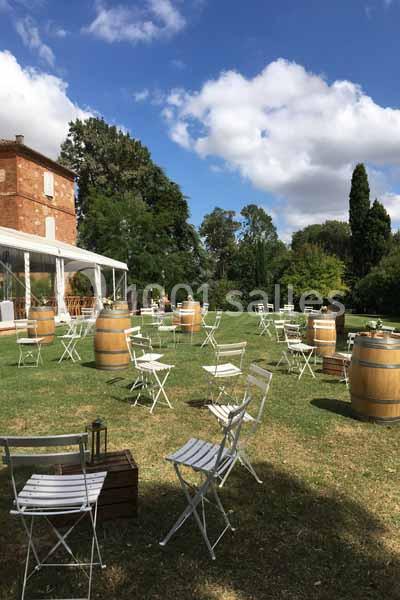 Chaises blanches et tonneaux disposés sur une pelouse devant un bâtiment en briques sous un ciel partiellement nuageux.