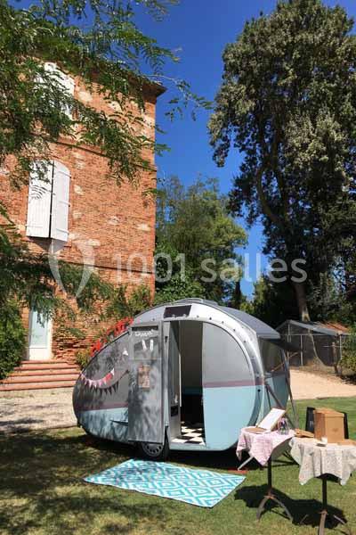 Caravane rétro stationnée sur une pelouse devant une maison en briques, avec des arbres et un ciel bleu en arrière-plan.