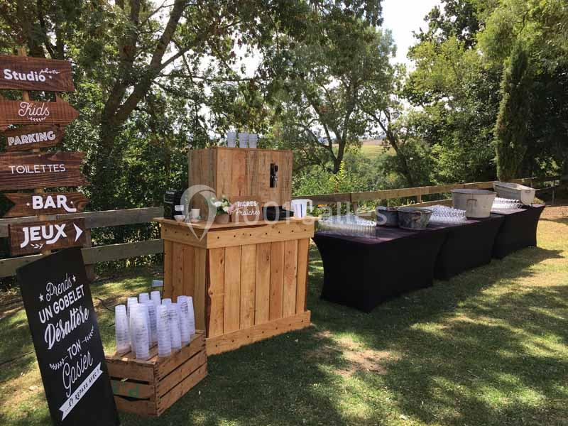 Stand en bois avec distributeur de boissons, gobelets et tables noires, installé en extérieur sous des arbres.