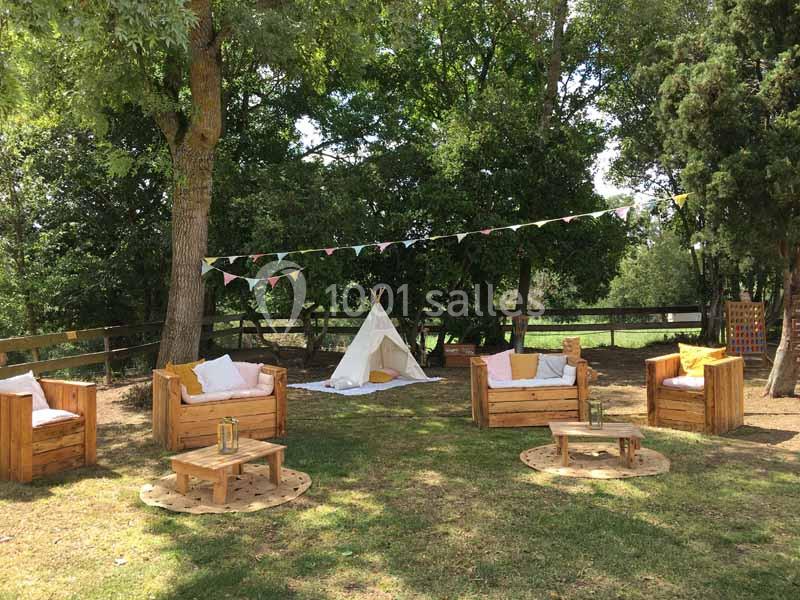 Mobilier en bois avec coussins disposé sur une pelouse ombragée, tipi blanc et guirlande de fanions en arrière-plan.