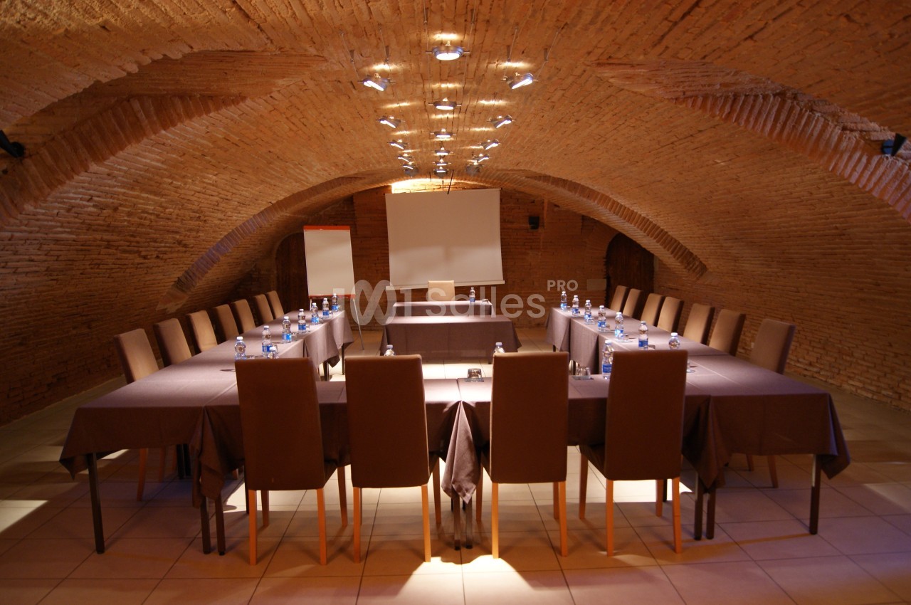 Salle voûtée en briques avec une grande table en U, chaises alignées, écran de projection et tableau blanc.
