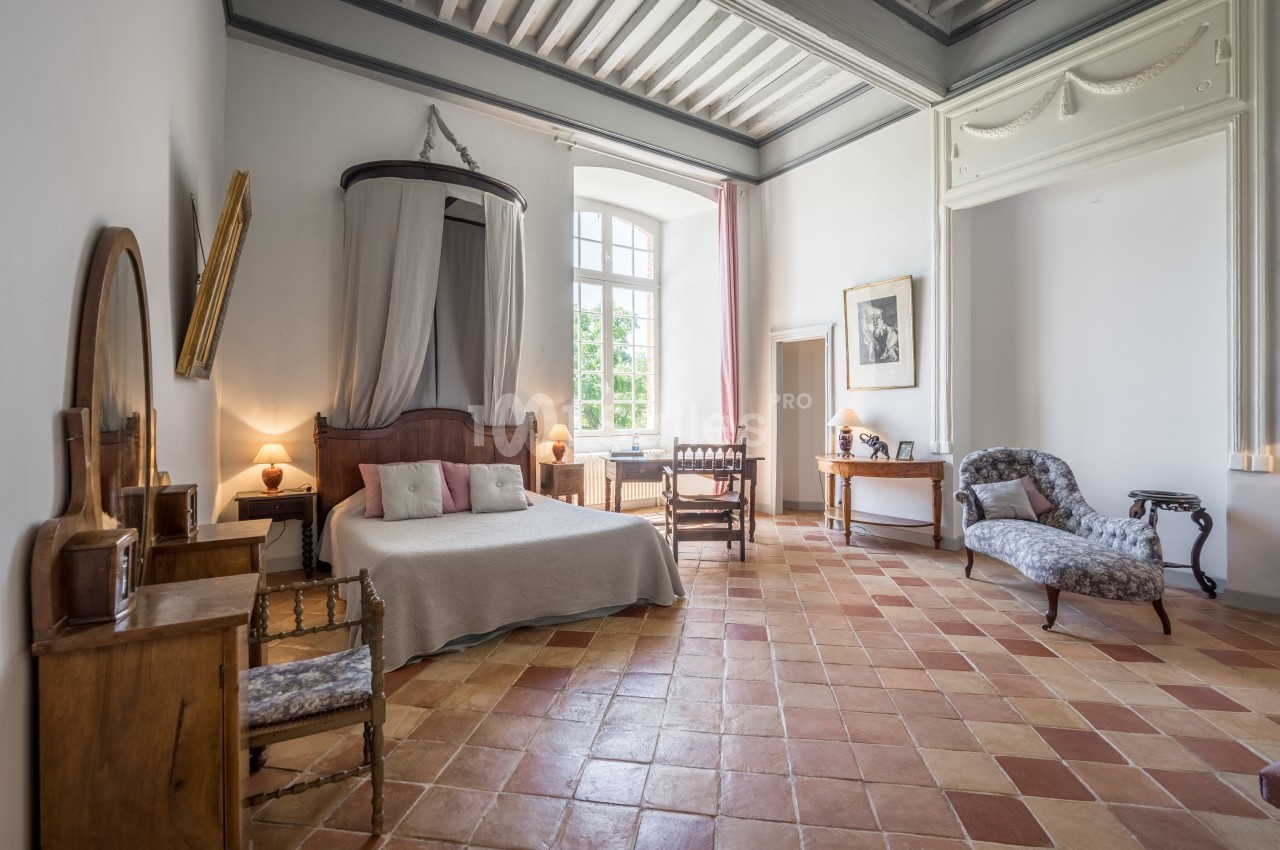 Chambre spacieuse avec lit à baldaquin, meubles anciens, sol en terre cuite et grande fenêtre laissant entrer la lumière.