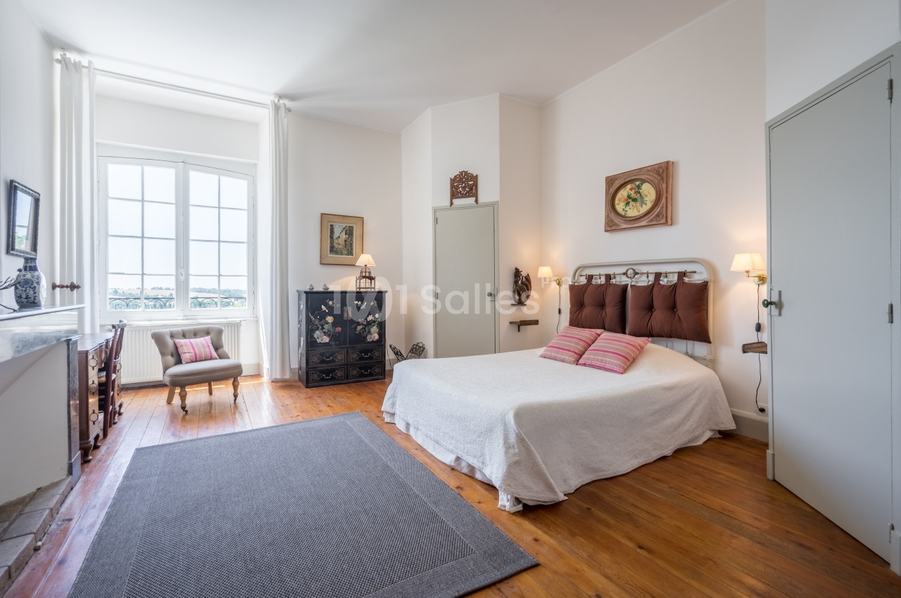 Chambre lumineuse avec lit double, parquet en bois, tapis gris, mobilier ancien et grande fenêtre donnant sur l'extérieur.