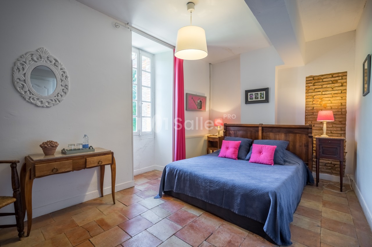 Chambre lumineuse avec lit double, coussins roses, bureau en bois, mur en briques et fenêtre laissant entrer la lumière.