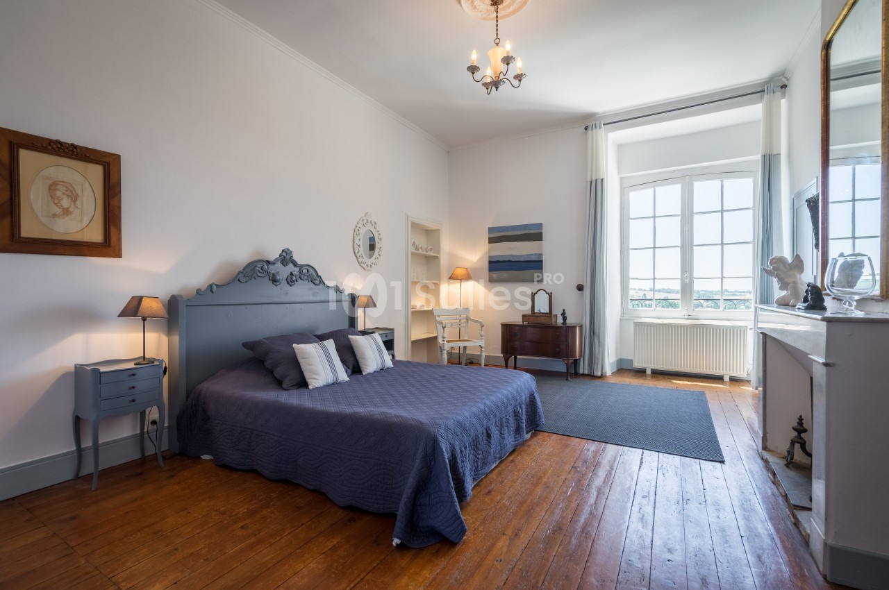 Chambre lumineuse avec lit double, parquet en bois, mobilier classique et grande fenêtre donnant sur l'extérieur.