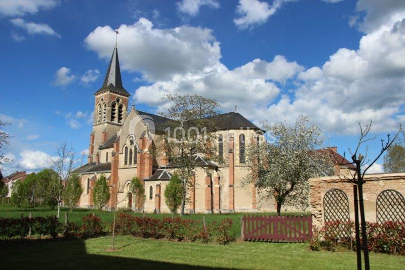 Église en pierre avec clocher, entourée d'arbres et de pelouses, sous un ciel bleu parsemé de nuages.