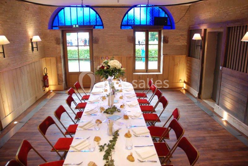 Salle décorée avec une grande table dressée pour un repas, entourée de chaises rouges, éclairée par des fenêtres et des…