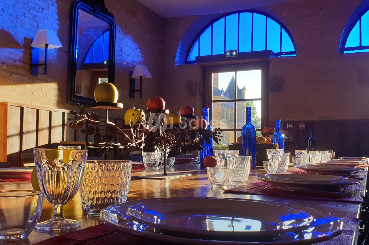 Table dressée avec vaisselle et décorations fruitées dans une salle lumineuse aux grandes fenêtres teintées de bleu.