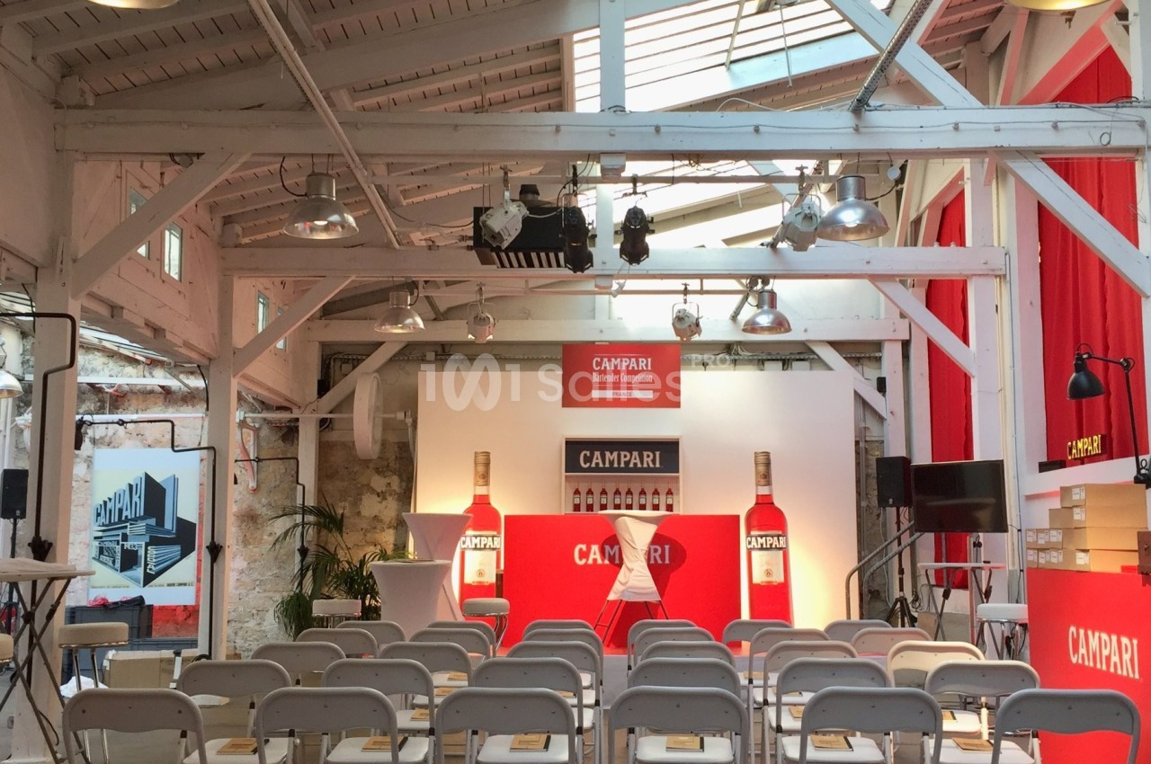 Salle lumineuse aménagée pour un événement, avec des chaises blanches alignées et une scène décorée aux couleurs de Campari.