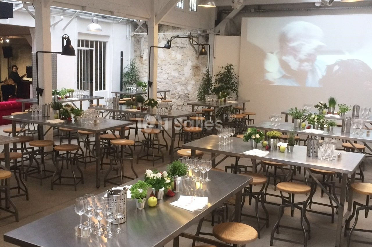 Salle lumineuse aménagée avec des tables en métal, des tabourets en bois et une décoration végétale.