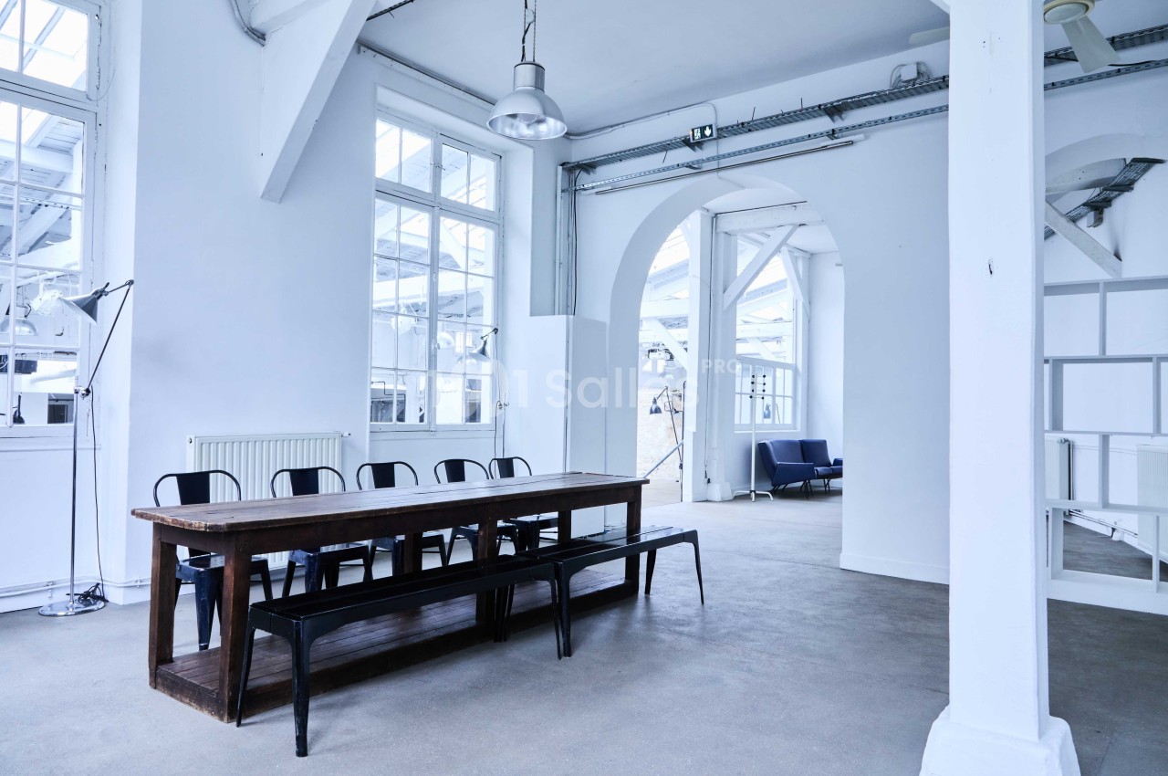 Salle lumineuse avec grande table en bois, chaises noires, murs blancs, fenêtres vitrées et mobilier minimaliste.