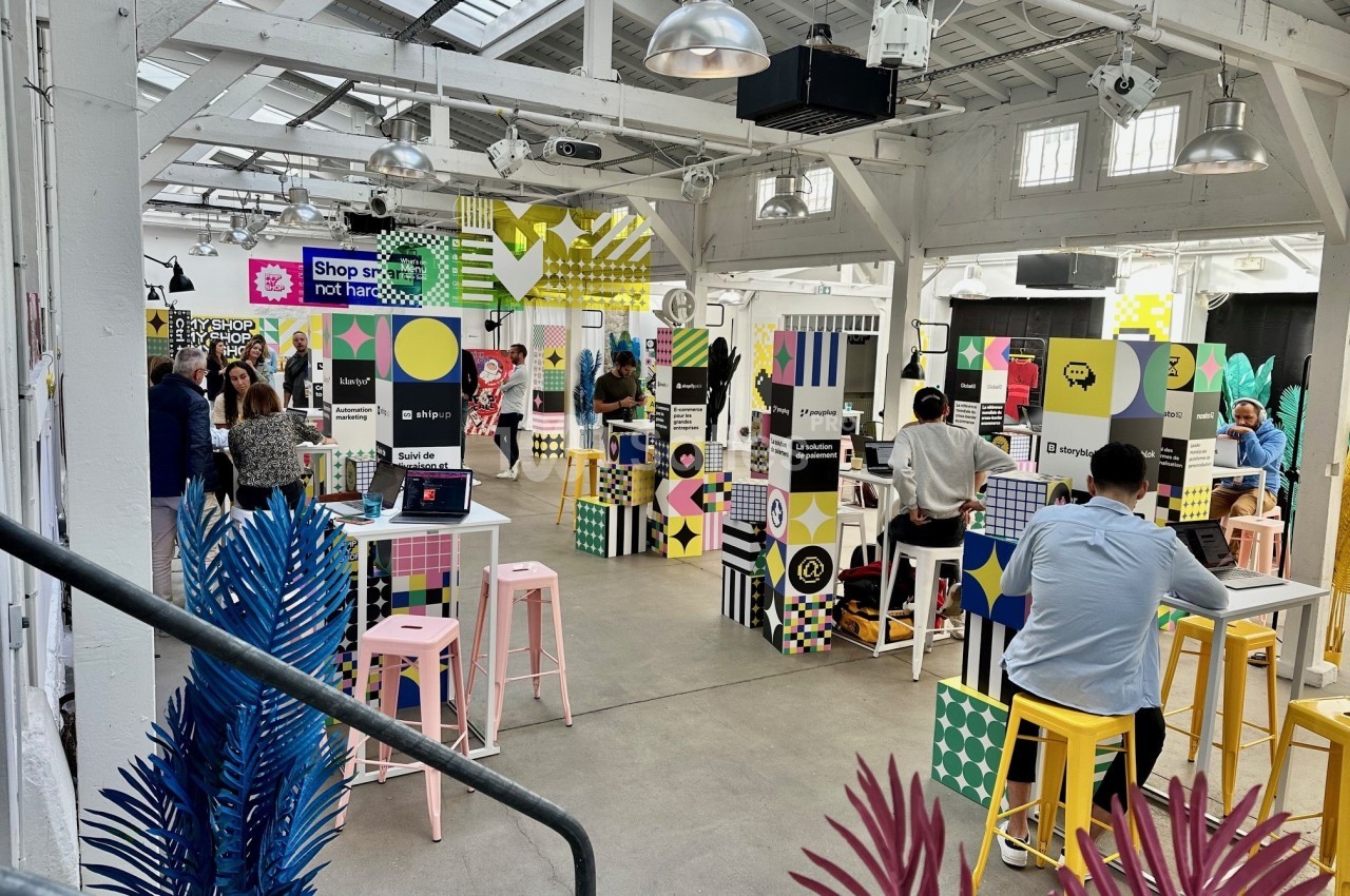 Espace lumineux avec des stands colorés, des visiteurs et une décoration moderne dans un bâtiment industriel.
