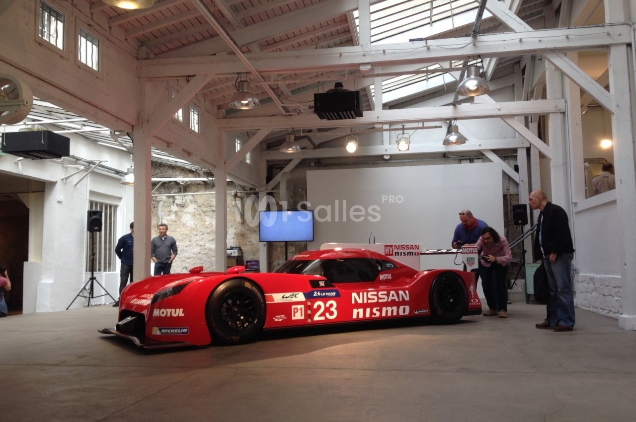 Voiture de course rouge exposée dans un espace intérieur avec des poutres blanches et quelques personnes autour.