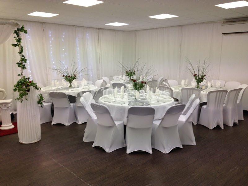Salle décorée avec des tables rondes dressées, nappes blanches, chaises houssées et arrangements floraux.