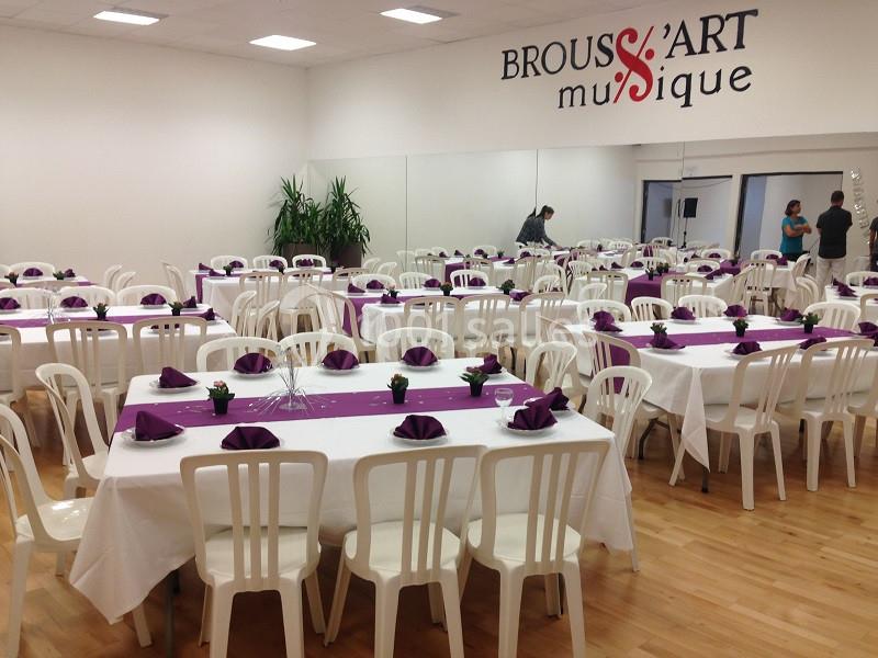 Salle aménagée avec des tables rectangulaires couvertes de nappes blanches et décorées de serviettes violettes.