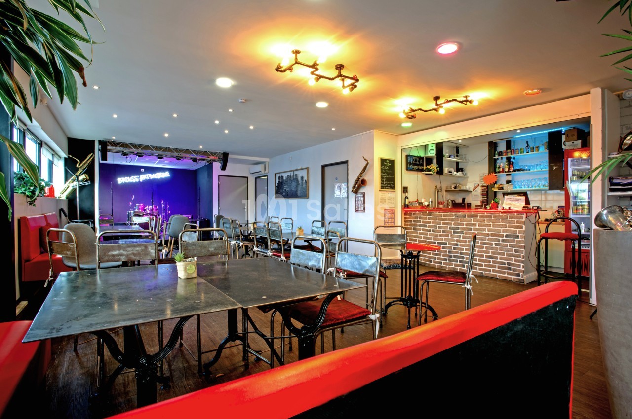 Salle de restaurant avec tables en métal, bar en briques, éclairage chaleureux et scène équipée en arrière-plan.