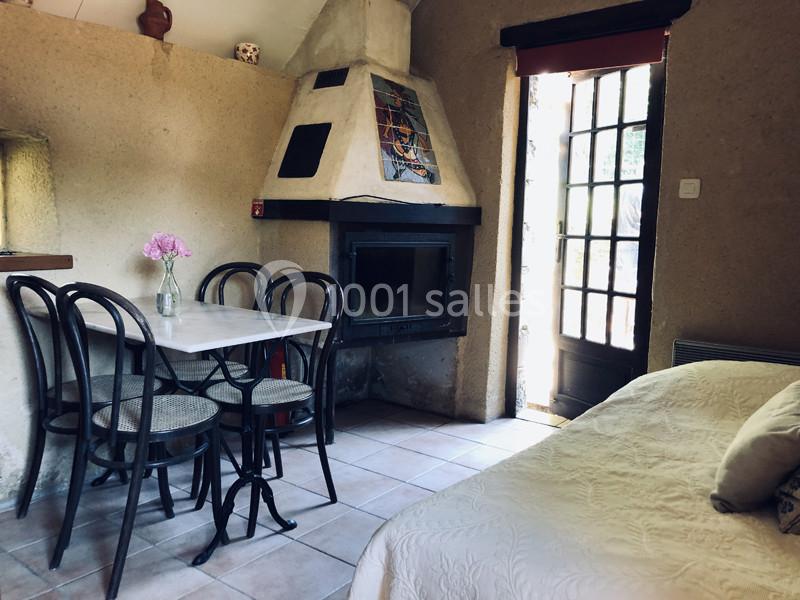 Salle à manger rustique avec table, chaises en bois, cheminée, lit et porte-fenêtre laissant entrer la lumière naturelle.