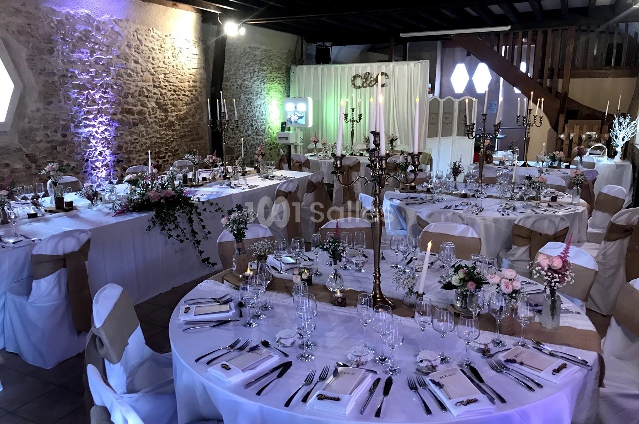 Salle de réception décorée pour un mariage, avec tables rondes dressées, chaises ornées de nœuds et éclairage tamisé.