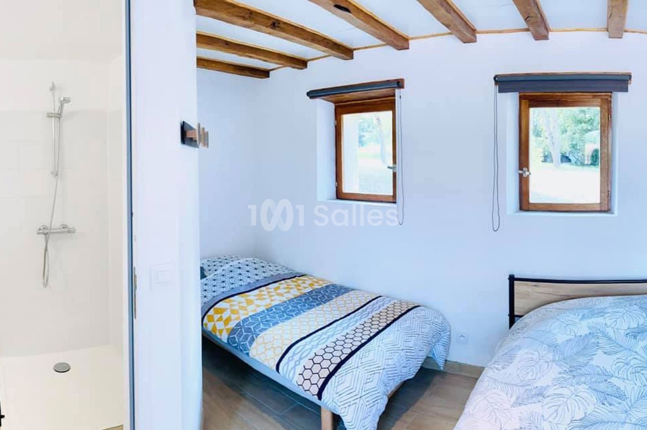 Deux lits simples dans une chambre lumineuse avec plafond en poutres apparentes, vue sur une douche et fenêtres.