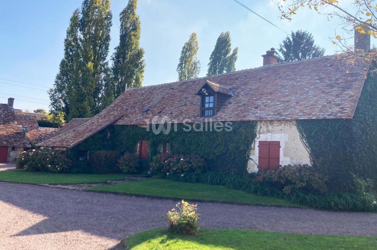 Maison ancienne en pierre avec toit en tuiles, façade recouverte de lierre, entourée d'arbres et d'un chemin en gravier.