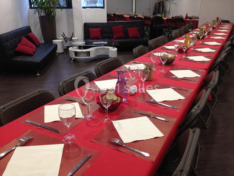 Salle aménagée pour un repas, avec longues tables rouges dressées, chaises pliantes et canapés en arrière-plan.