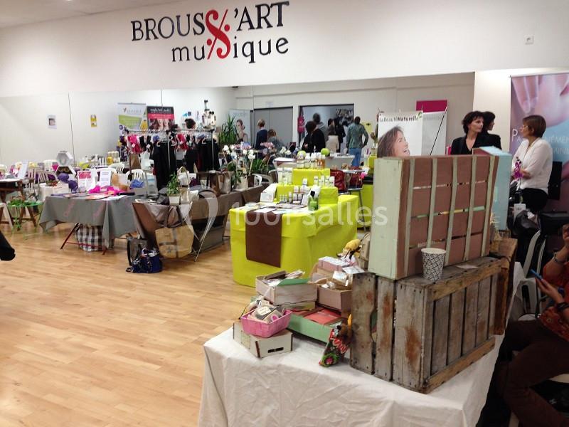 Salle d'exposition avec stands de produits artisanaux, cosmétiques et décorations, sur un sol en parquet clair.