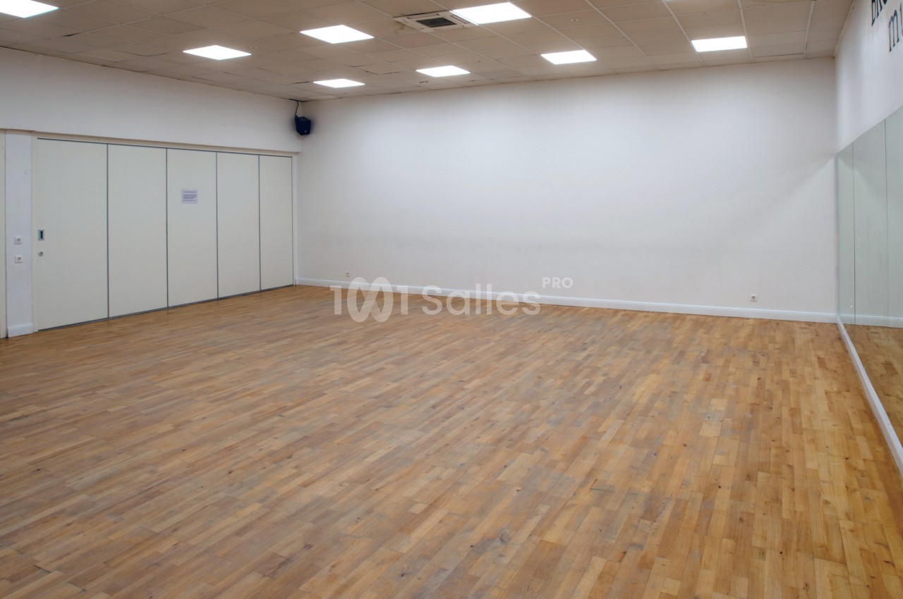 Salle vide avec parquet en bois, murs blancs, plafond à dalles lumineuses et grand miroir sur un côté.