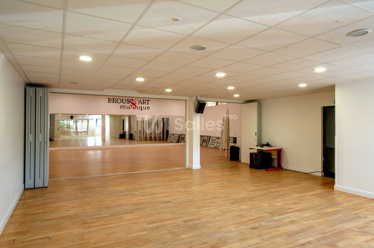 Salle spacieuse avec parquet, miroirs muraux, chaises empilées et éclairage au plafond.