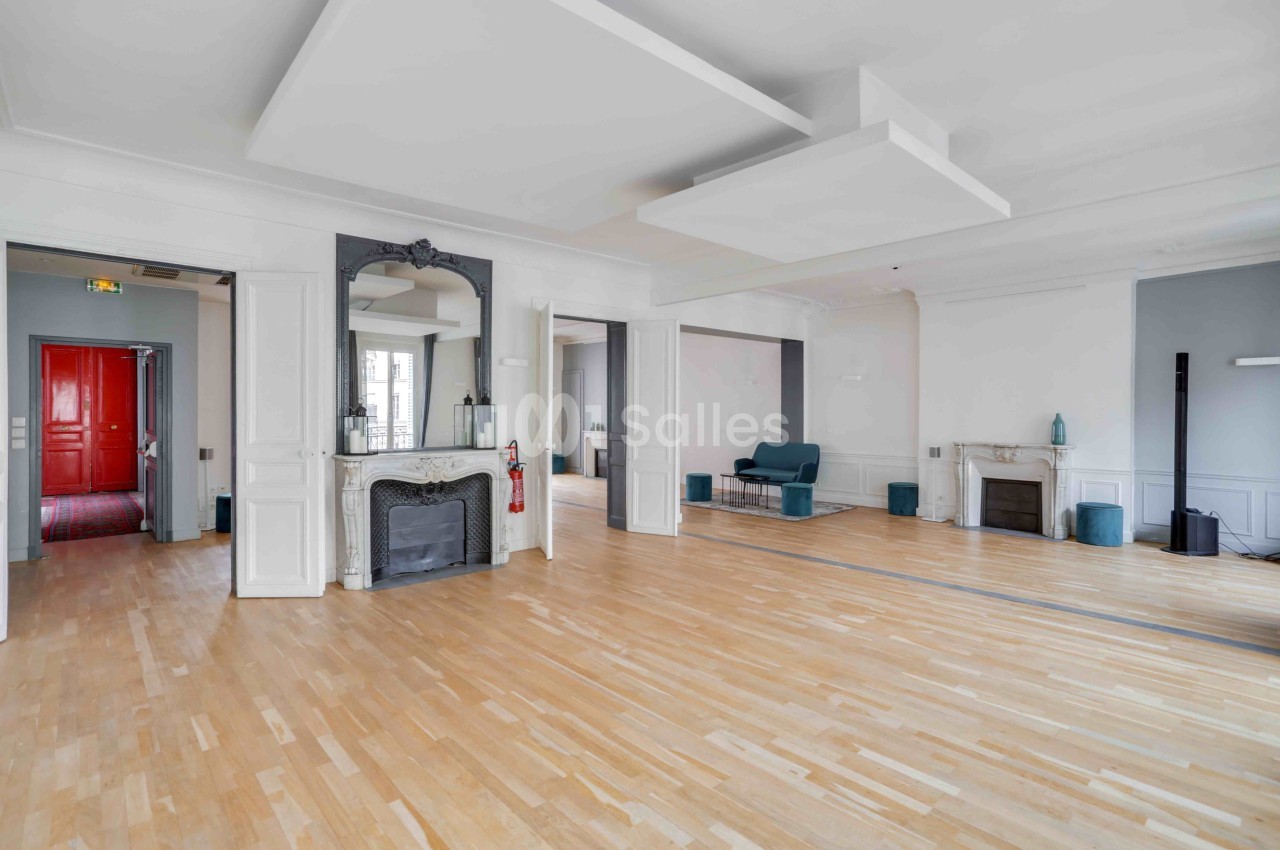 Salle spacieuse avec parquet clair, cheminée, miroir, murs blancs et une porte rouge visible au fond.