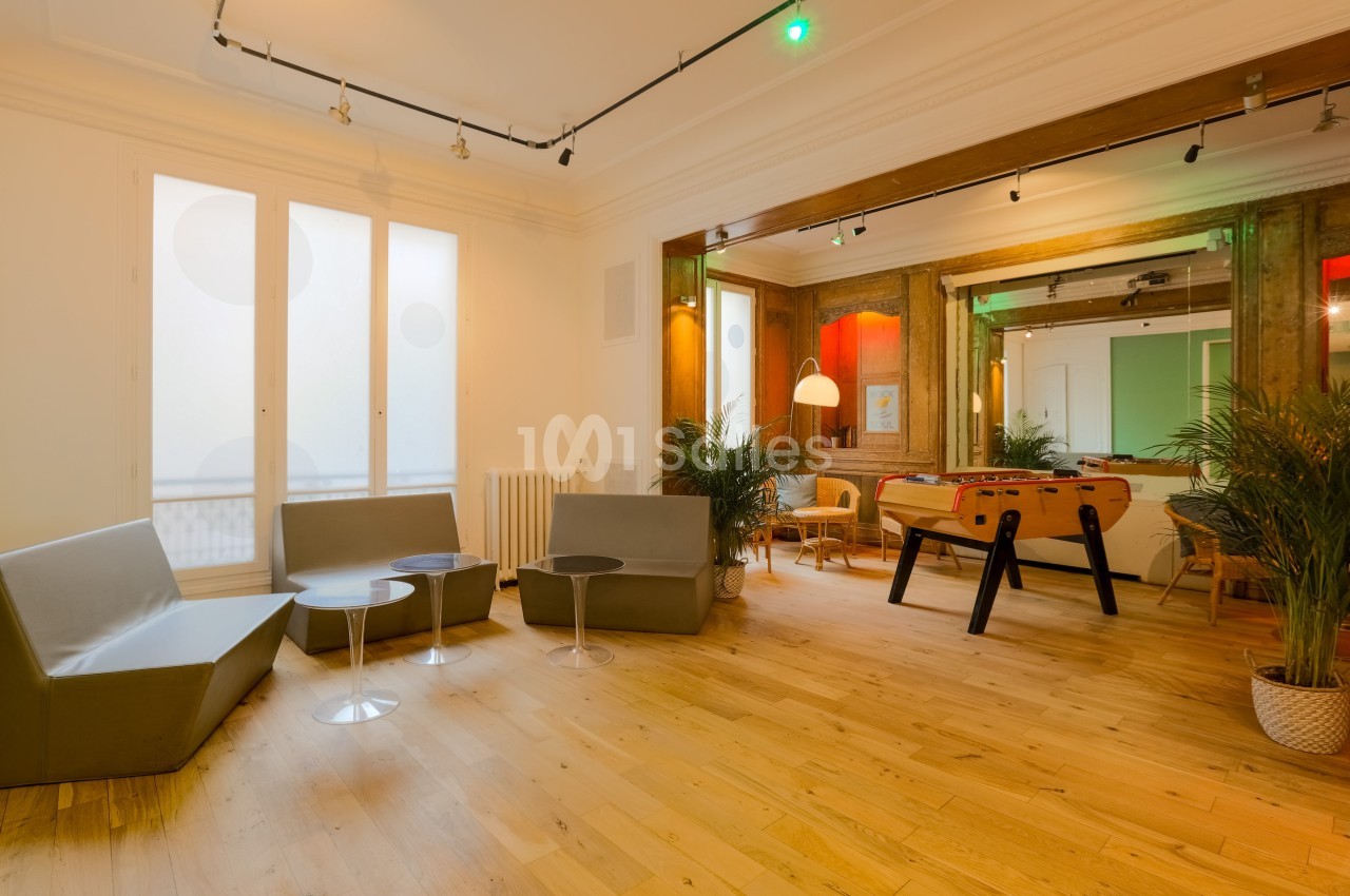 Salle lumineuse avec parquet, canapés gris, baby-foot, plantes et grandes fenêtres laissant entrer la lumière naturelle.